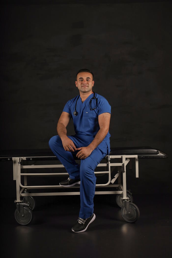 portfolio-02 Male doctor in blue scrub suit seated on a stretcher with a stethoscope, smiling on a dark studio background.